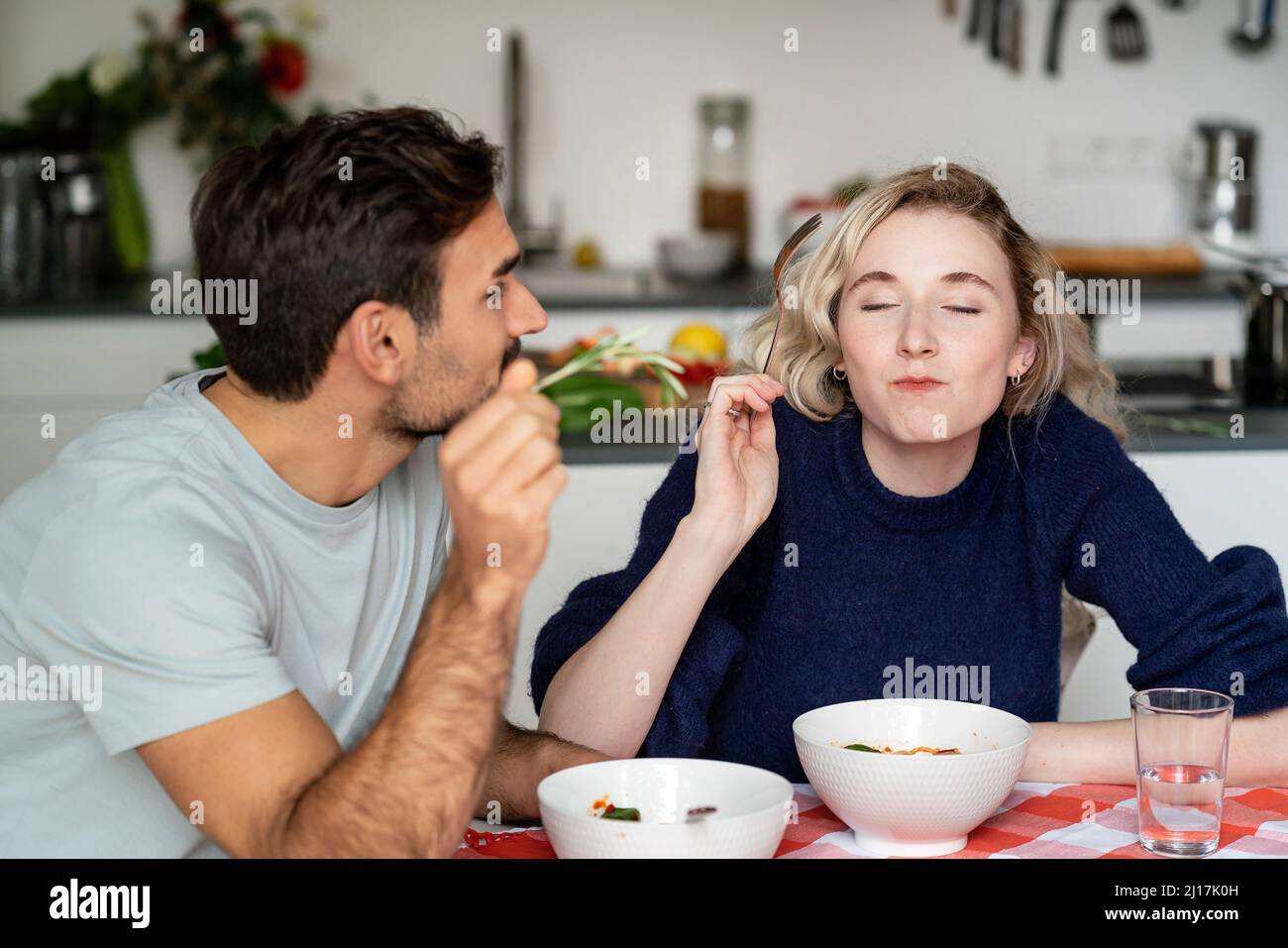 Junger Mann, der die Freundin beim Essen ansah, mit geschlossenen Augen am Tisch sitzend Stockfoto