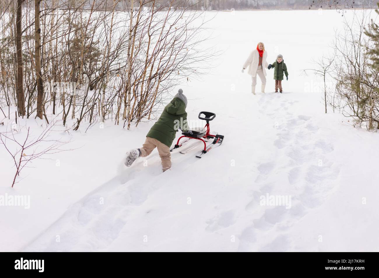 Frau mit Sohn, der im Winter den Jungen ansieht, der im Schnee Rodel schiebt Stockfoto