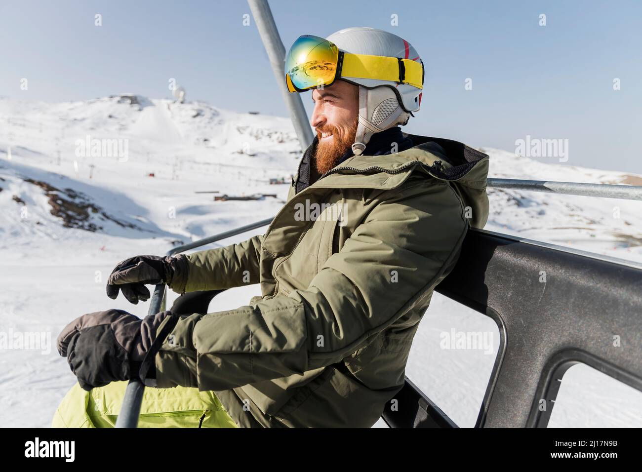 Lächelnder junger Mann in einer Jacke mit Skibrille und Helm, der auf dem Skilift sitzt Stockfoto