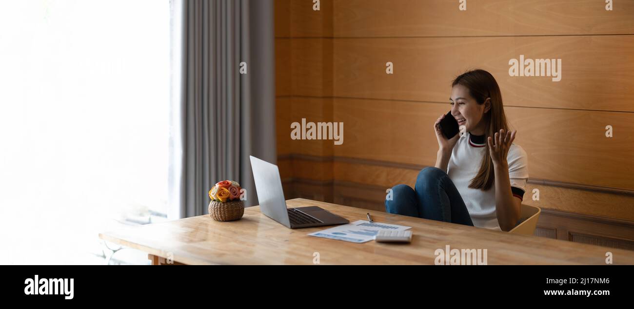 Glückliche junge asiatische Frau, die auf dem Handy spricht und lächelt, während sie zu Hause mit Laptop und Papierkram arbeitet Stockfoto