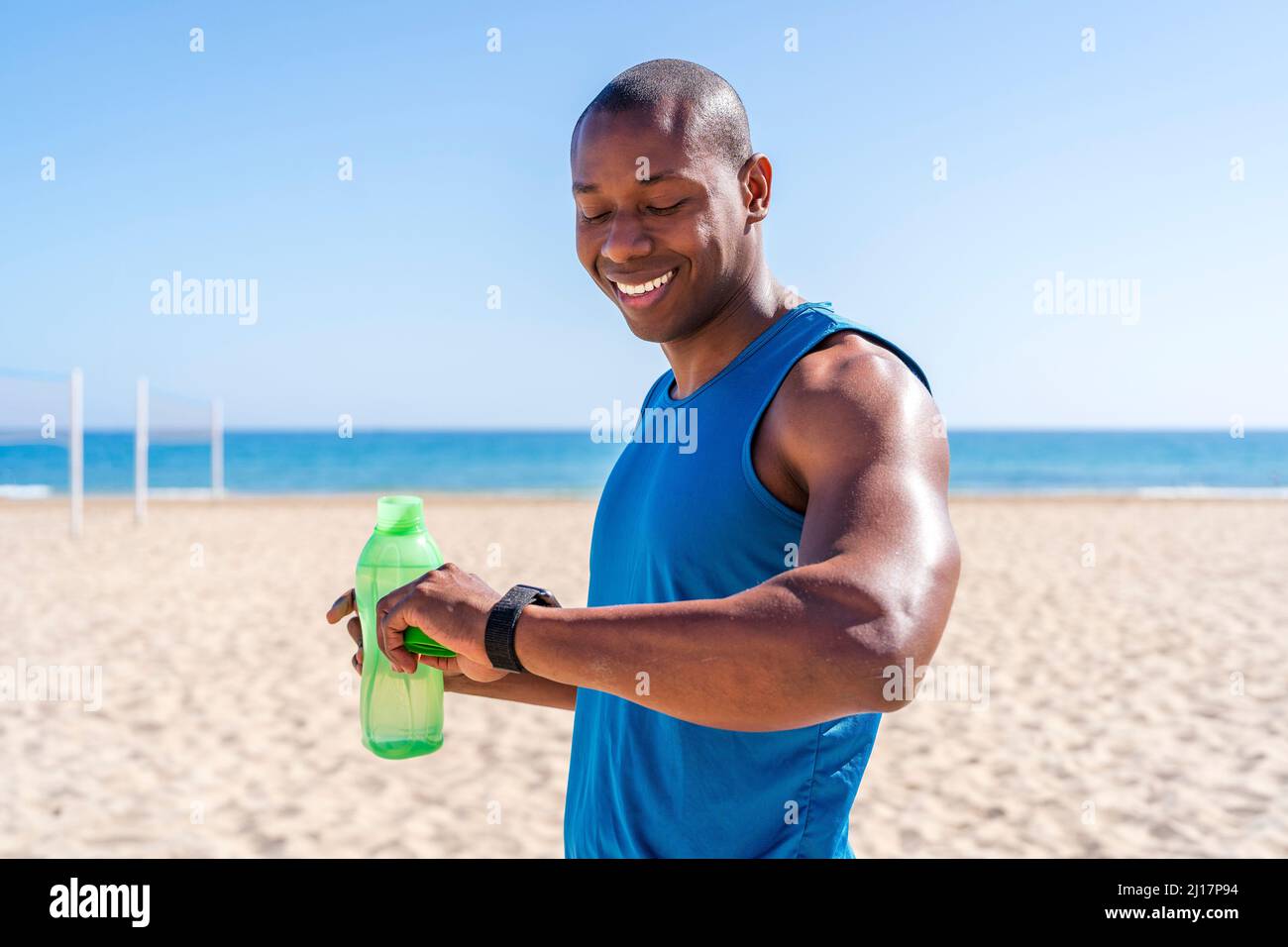 Lächelnder Mann mit Wasserflasche, der die Zeit am Strand an einem sonnigen Tag überprüft Stockfoto