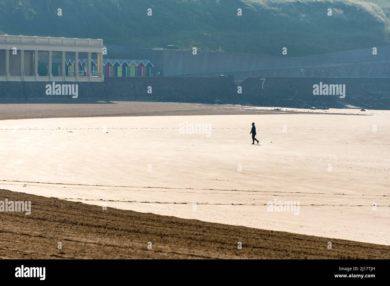 Eine einsame Person geht am frühen Morgen an einem sonnigen Sommertag über den Sandstrand von Whitmore Bay, Barry Island. Stockfoto
