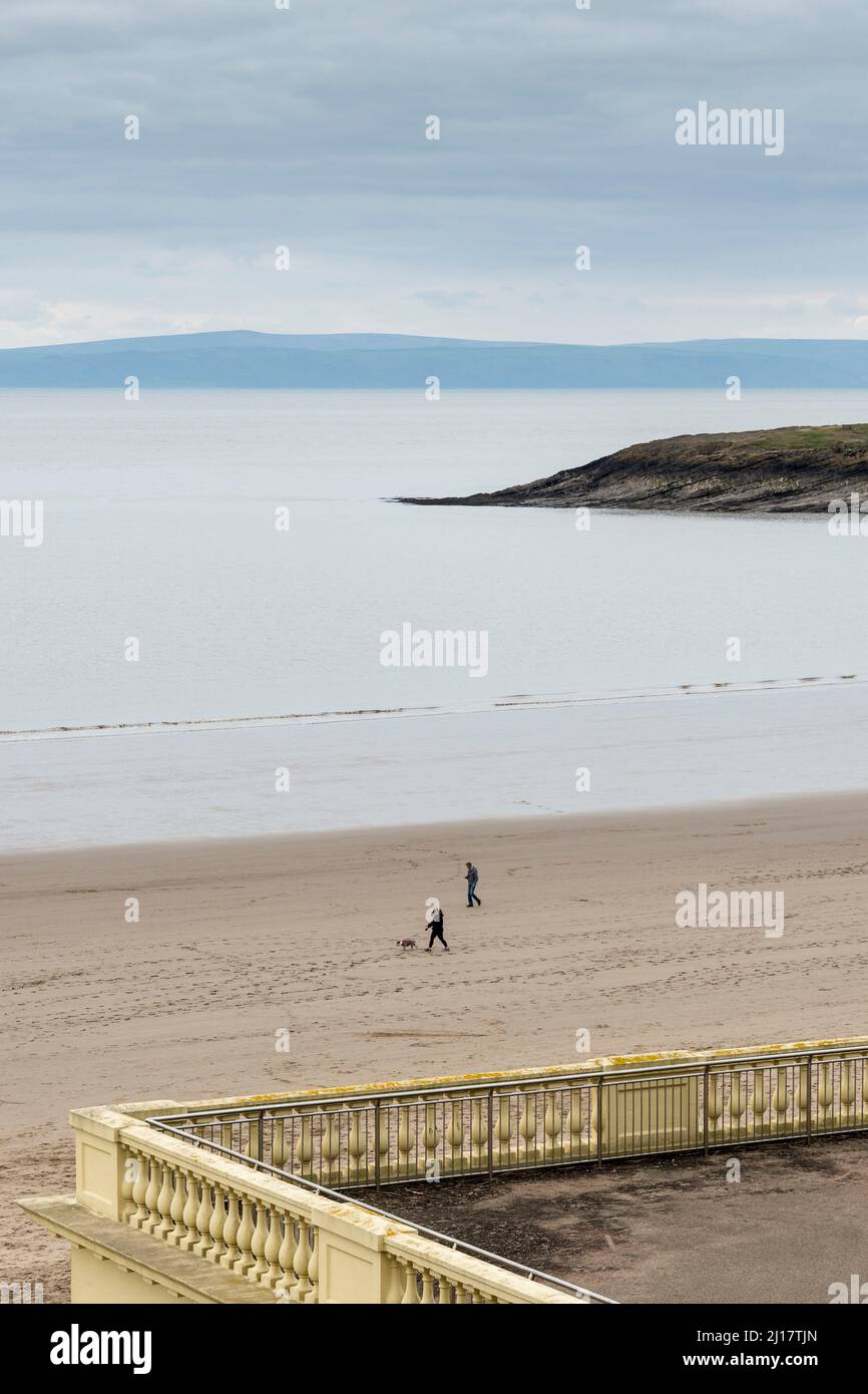 Zwei Personen, einer mit einem Hund, spazieren an einem ruhigen, bewölkten Tag am Sandstrand Barry Island entlang. Exmoor liegt am Horizont über den Bristol Channel. Stockfoto