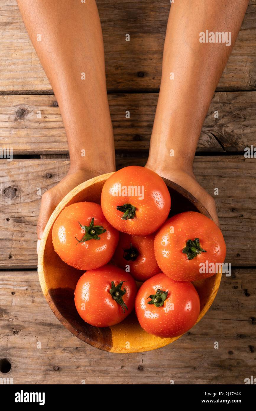 Ansicht von oben auf die Hände des Mannes, der eine Schüssel voller frischer Tomaten in einer Schüssel auf einem Holztisch hielt Stockfoto