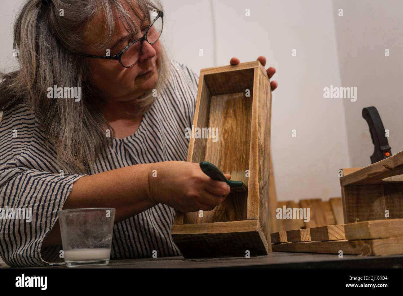 Weißhaarige Frau, die in ihrer Werkstatt für recyceltes Holz mit verschiedenen Werkzeugen und ökologischen Materialien arbeitet.Holz lackieren Stockfoto