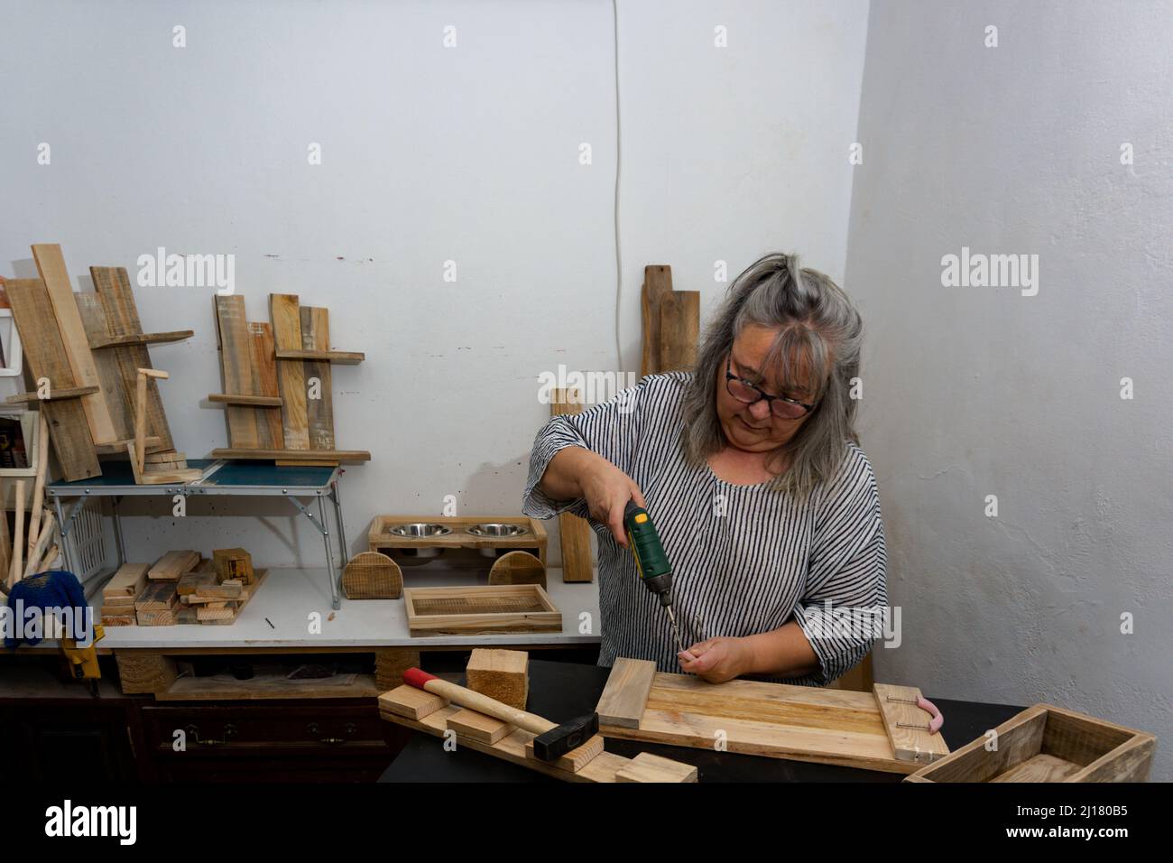 Weißhaarige Frau, die in ihrer Werkstatt aus recyceltem Holz mit verschiedenen Werkzeugen und ökologischen Materialien arbeitet.mit einem elektrischen Schraubendreher Stockfoto
