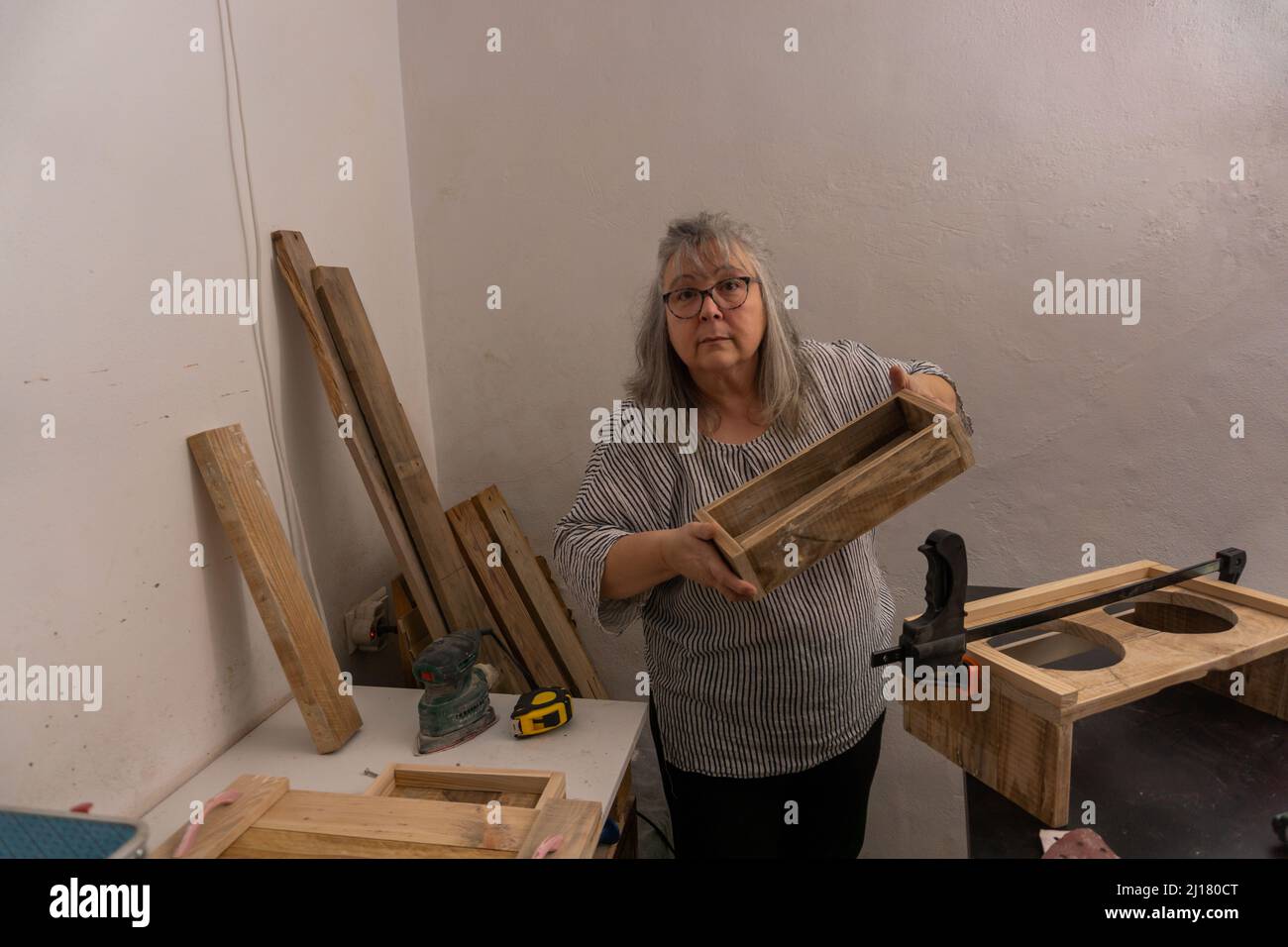 Weißhaarige Frau, die in ihrer Werkstatt aus recyceltem Holz mit verschiedenen Werkzeugen und ökologischen Materialien arbeitet und ihre Arbeit zeigt Stockfoto
