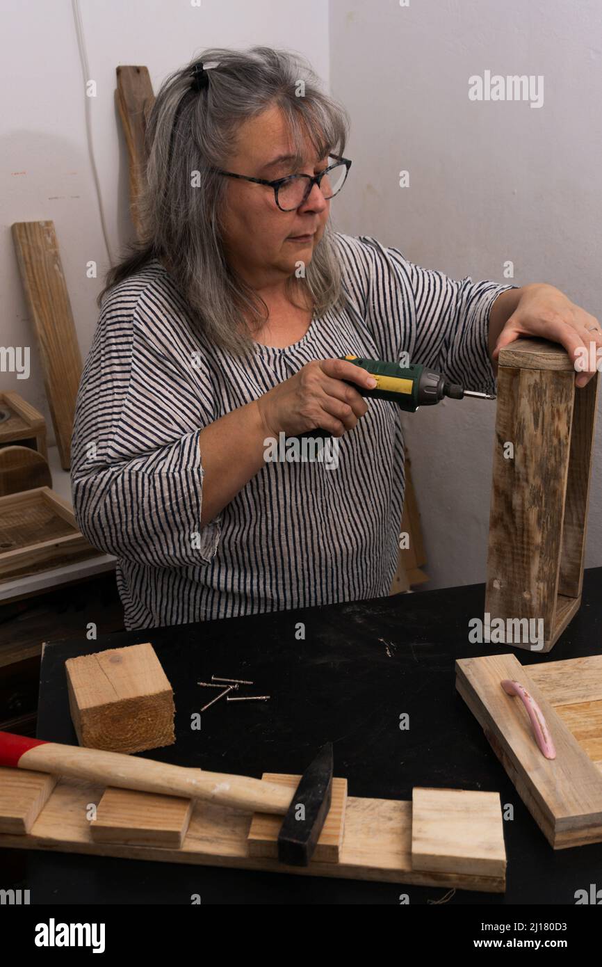 Weißhaarige Frau, die in ihrer Werkstatt aus recyceltem Holz mit verschiedenen Werkzeugen und ökologischen Materialien arbeitet.mit einem elektrischen Schraubendreher Stockfoto