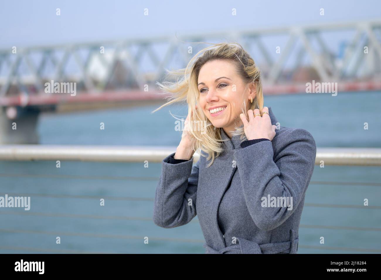 Attraktive Frau, die an einem windigen Tag an einer Uferpromenade steht und ihre langen blonden Haare mit einem Lächeln anklammert Stockfoto