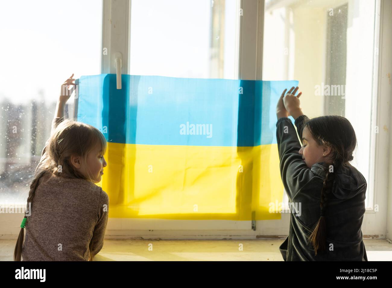 Kleine Mädchen mit ukrainischer Flagge vor einem Fenster. Die kleinen Mädchen winken die Nationalflagge, während sie für den Frieden beten Stockfoto