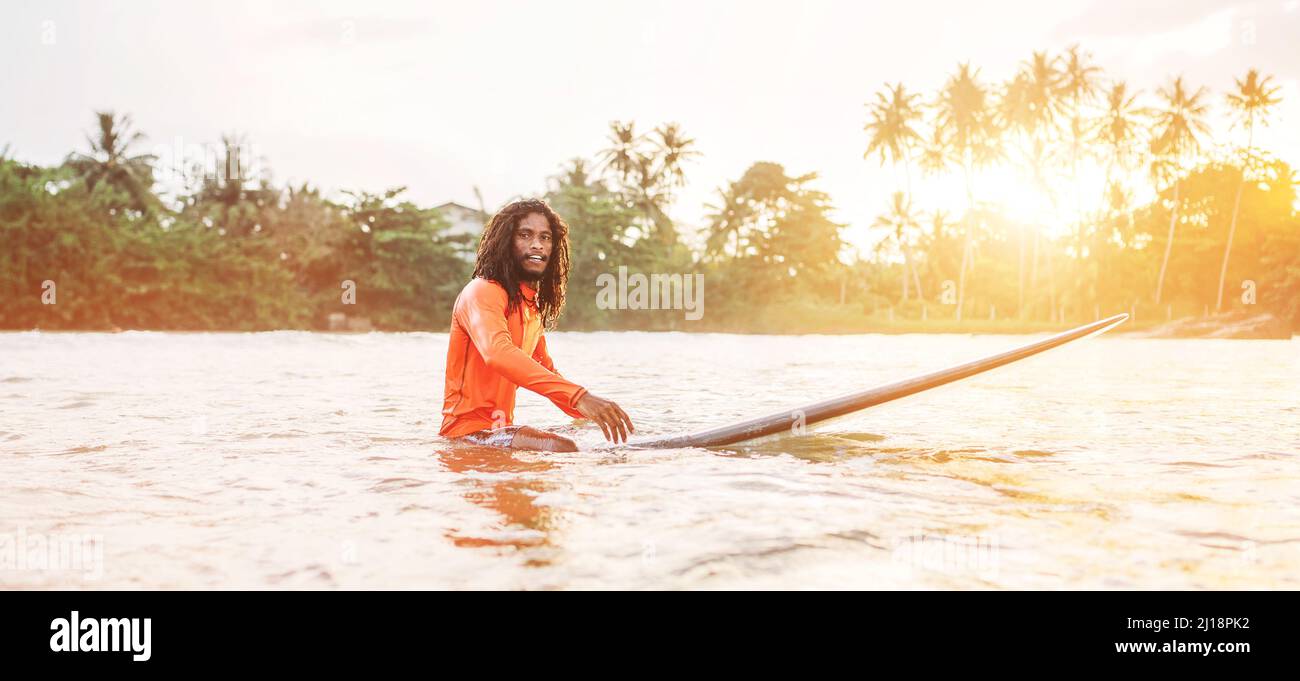 Schwarzer langhaariger Teenager-Mann, der auf einem langen Surfbrett schwimmt und auf eine Welle wartet, die zum Surfen bereit ist, mit Palmenhain-Sonnenstrahlen. Extremwassersport a Stockfoto
