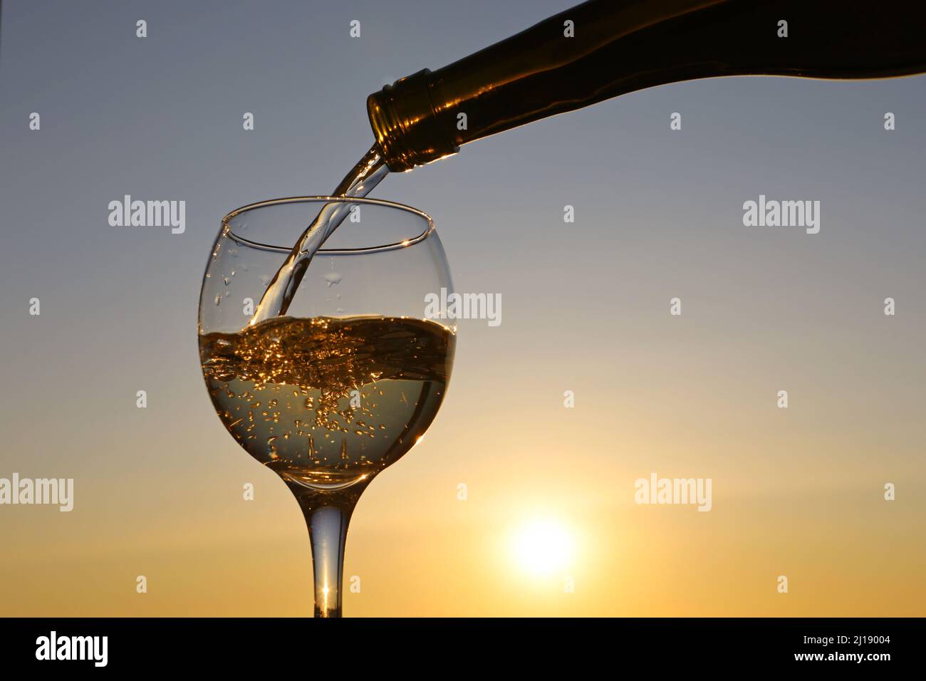 Gießen Sie Weißwein aus einer Flasche in das Glas auf Sonnenuntergang Himmel Hintergrund. Feier, romantisches Abendessen mit Alkoholgetränk Stockfoto