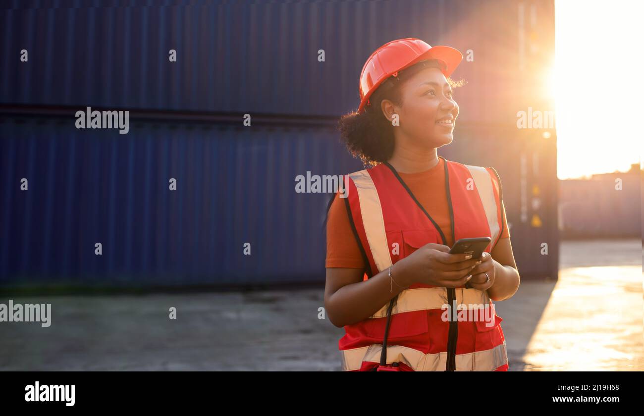 Porträt einer glücklichen, lächelnden afroamerikanischen Ingenieurin, die Sicherheitsweste und Helm trägt, auf logistischer Transportfracht stehend und mit dem Mobiltelefon unterwegs ist Stockfoto