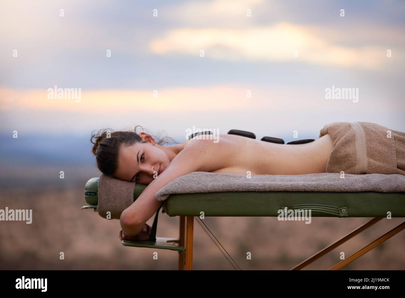 Heiße Steine werden diese müden Knochen ausheben. Porträt einer jungen Frau, die draußen auf einem Massageliege eine Behandlung mit heißen Steinen erhält. Stockfoto