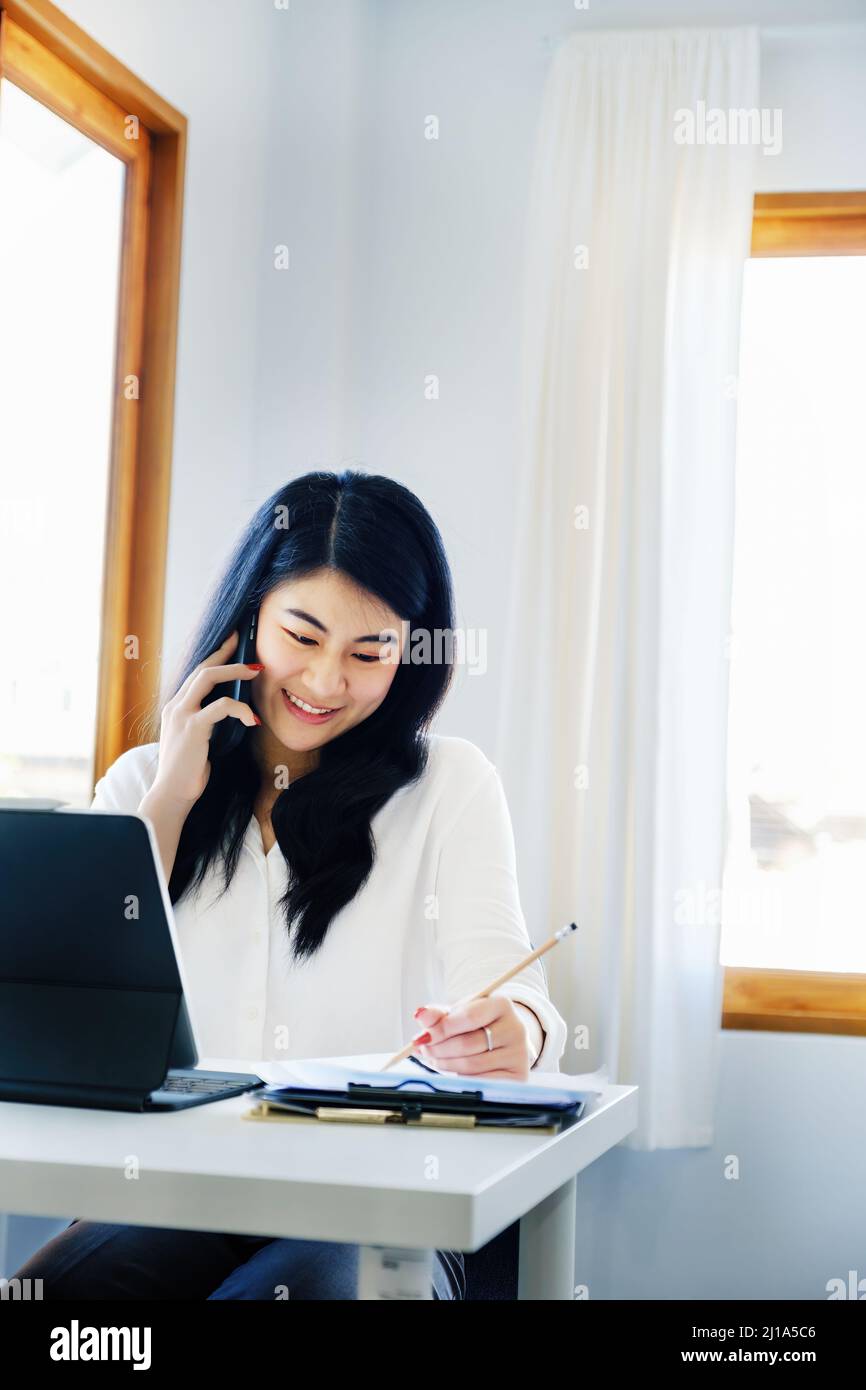 Geschäftsbeziehungen, ist ein asiatischer Sekretär des Unternehmens am Telefon mit Kunden kommt mit Stift in Kontakt, um die Zeit aufzuschreiben, um die zusammenzufassen Stockfoto