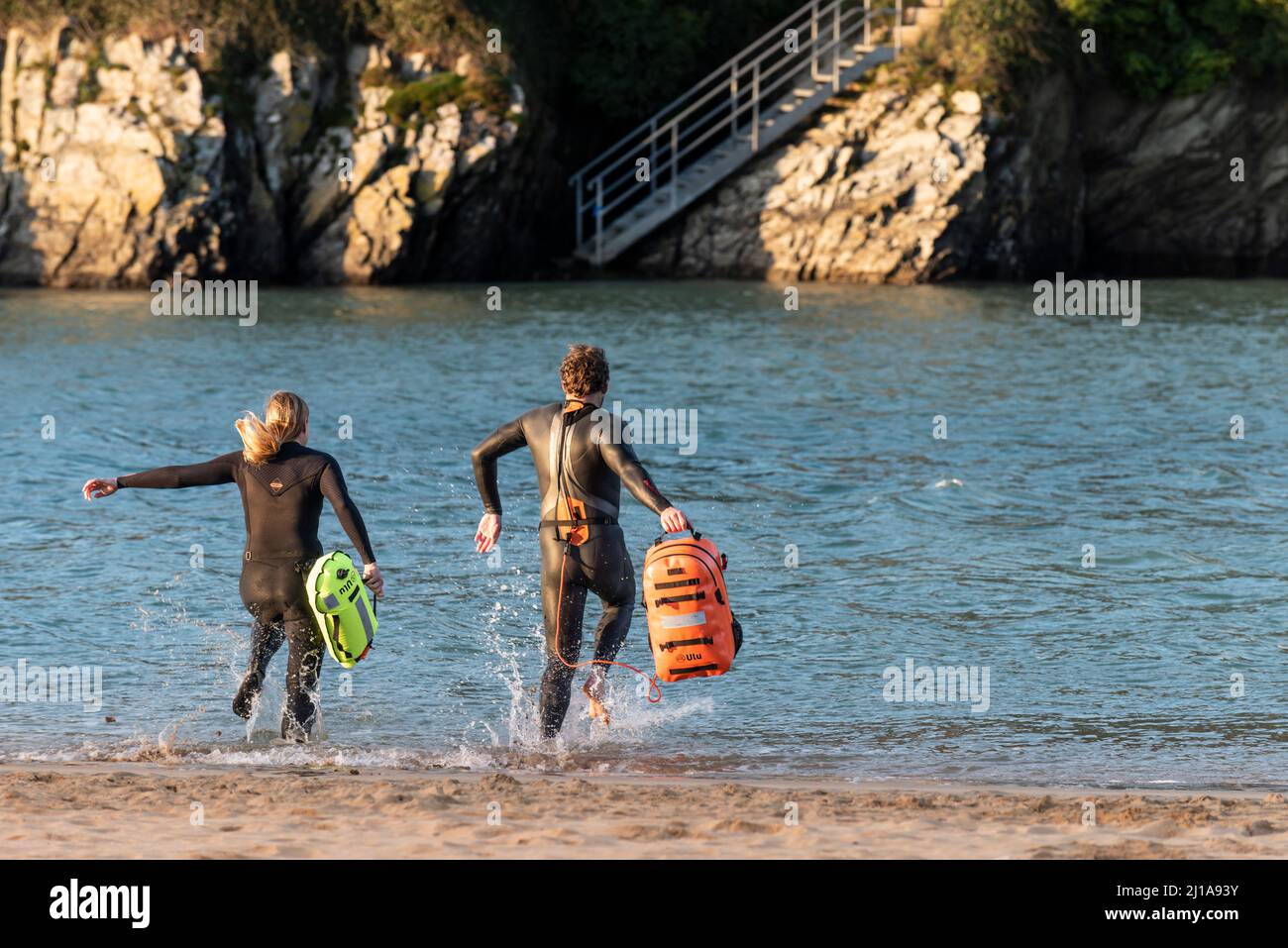 Wilde Schwimmer in Neoprenanzügen und mit fluurescent ULU Aufblasbare Dry Bags, die zum Gannel River am Crantock Beach in Newquay in Cornwal laufen Stockfoto