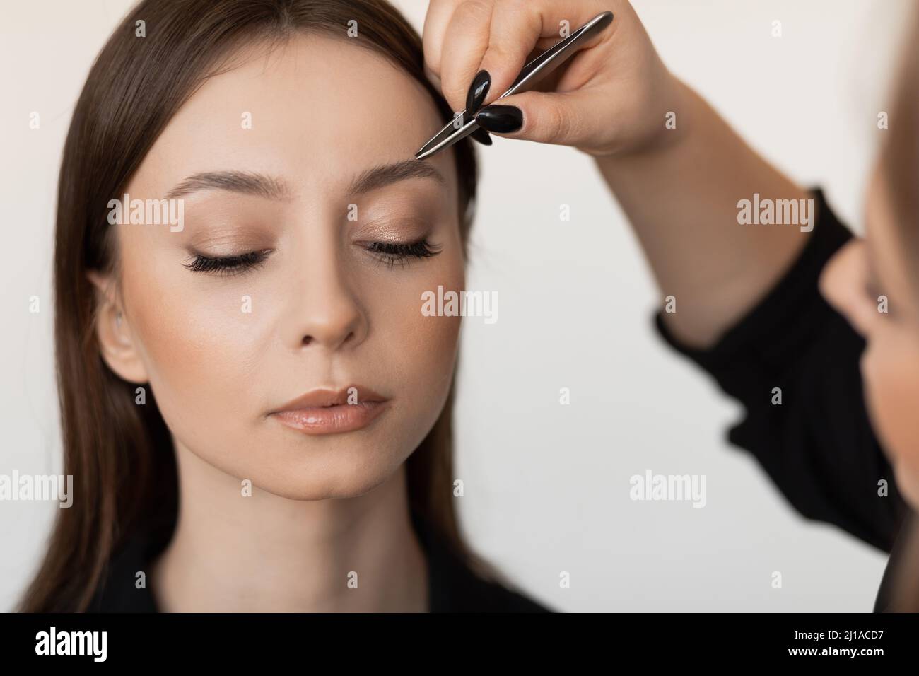 Nahaufnahme einer jungen perfekten Frau mit langen dunklen Haaren, die Augenbrauen zettelt, von einem professionellen Make-up-Künstler in schwarzer Kleidung. Stockfoto