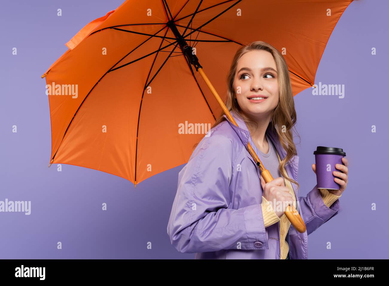 Fröhliche junge Frau im Trenchcoat, die Papierbecher hält und unter einem orangefarbenen Regenschirm auf lila isoliert steht Stockfoto