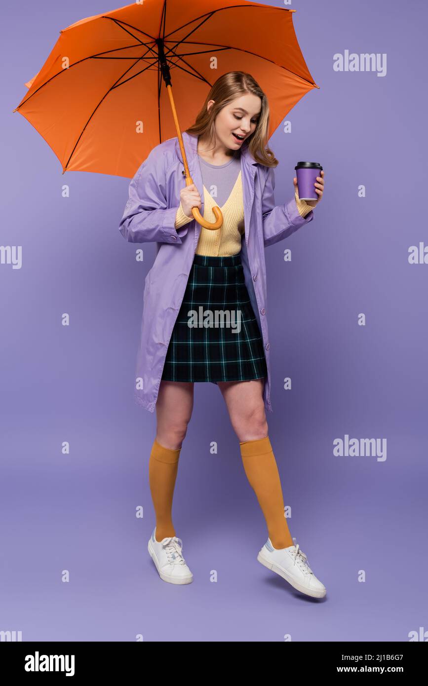 Die ganze Länge der staunenden jungen Frau in einem Trenchcoat hält einen Pappbecher und läuft unter einem orangefarbenen Regenschirm auf lila Stockfoto