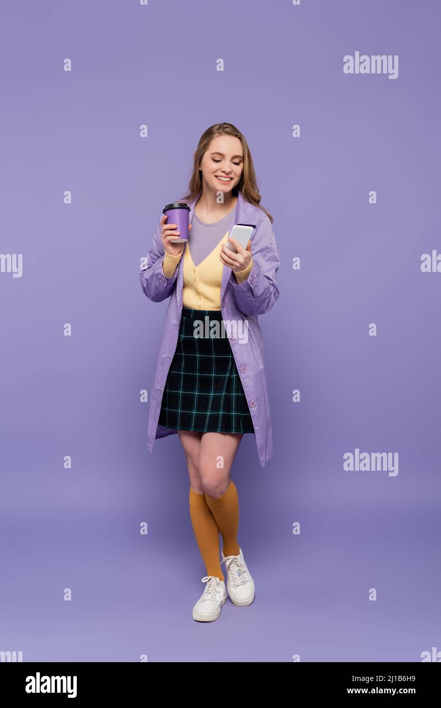 Volle Länge der glücklichen jungen Frau in Trenchcoat mit Smartphone und halten Papierbecher auf lila Stockfoto
