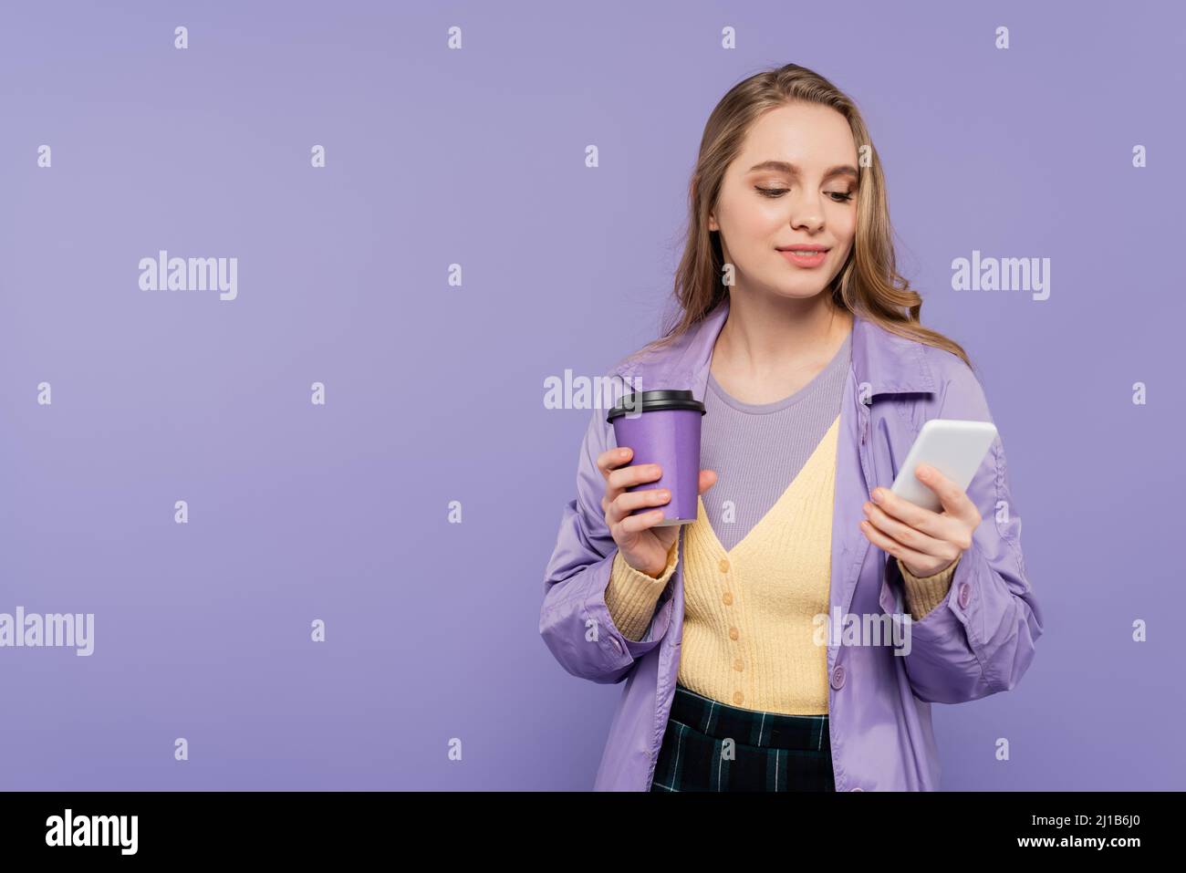 Glückliche junge Frau in Trenchcoat mit Smartphone und halten Papierbecher isoliert auf lila Stockfoto