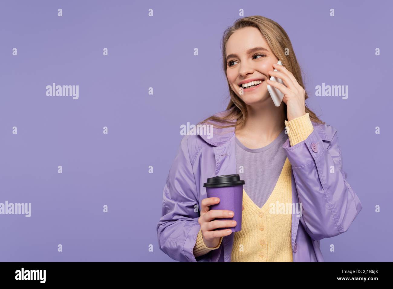 Lächelnde junge Frau im Trenchcoat, die auf dem Smartphone spricht und Papierbecher isoliert auf Violett hält Stockfoto