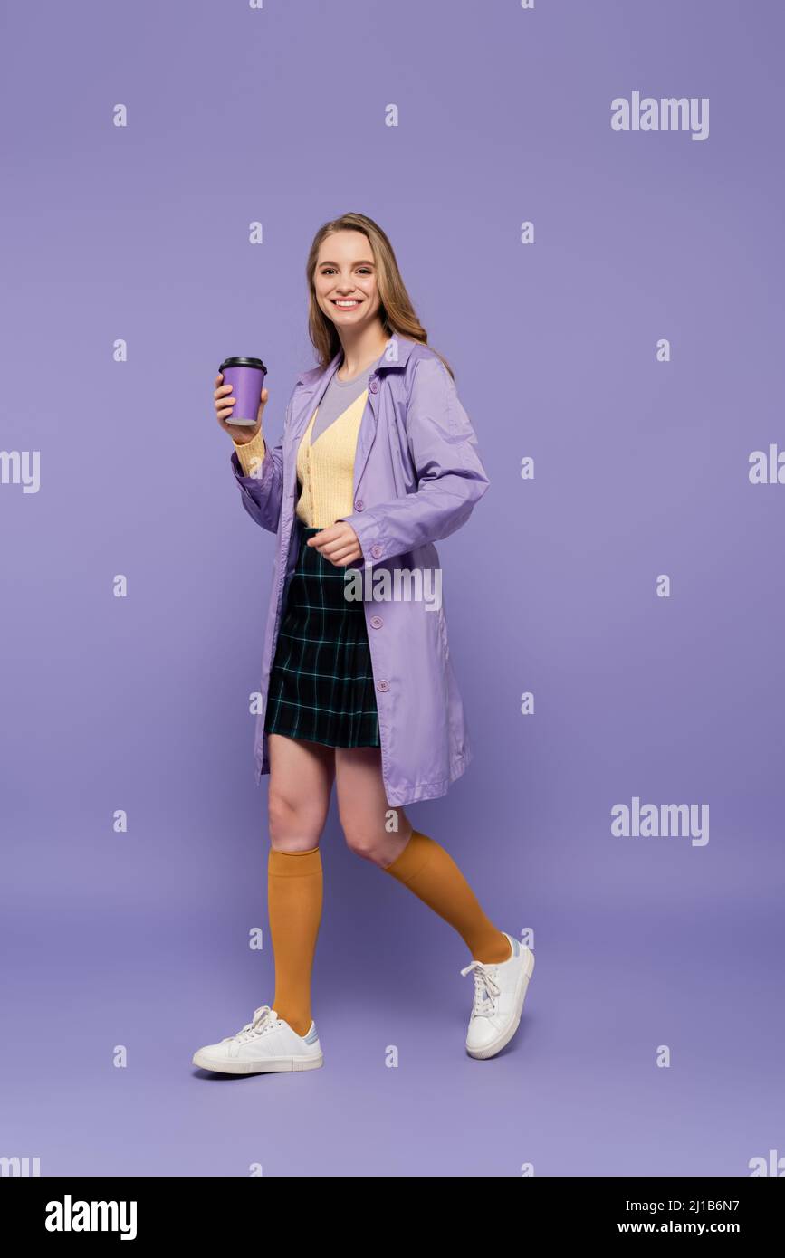 Die ganze Länge der glücklichen jungen Frau im Trenchcoat hält Papierbecher und geht auf lila Stockfoto