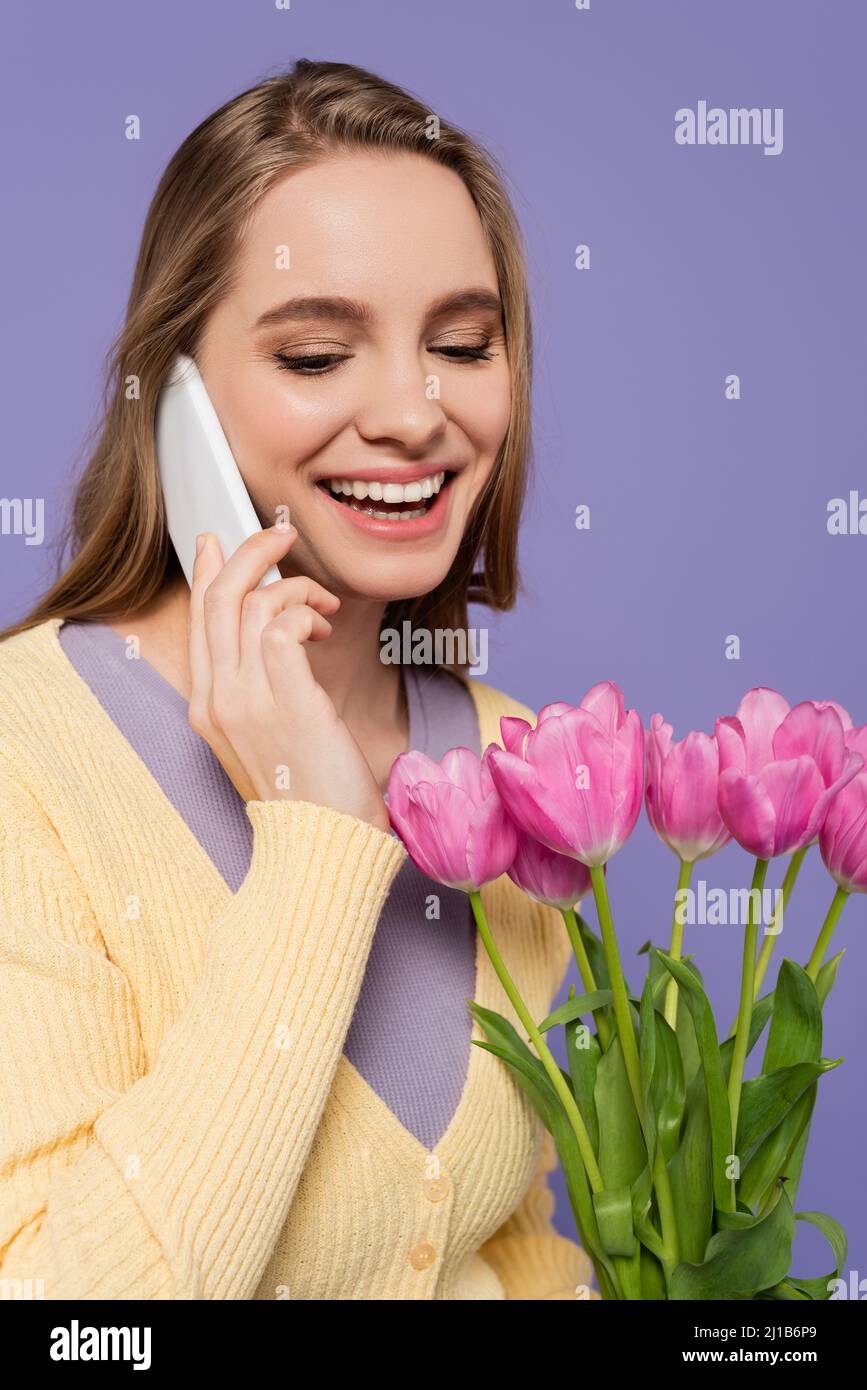Fröhliche junge Frau, die rosa Tulpen hält und auf dem Smartphone isoliert auf lila spricht Stockfoto