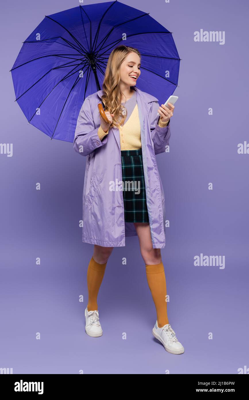 Volle Länge der glücklichen Frau zu Fuß mit Smartphone unter Regenschirm auf lila Stockfoto
