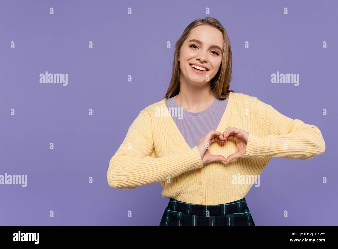 Fröhliche junge Frau mit Herzzeichen und Händen auf lila isoliert Stockfoto