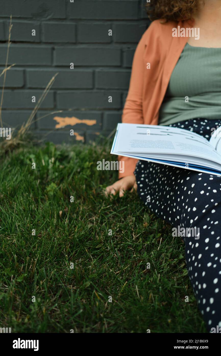 Junger Erwachsener Millennial mit lockigem Haar sitzt auf Gras. Student liest Buch. Lebensstil und Körper positiv. Stockfoto