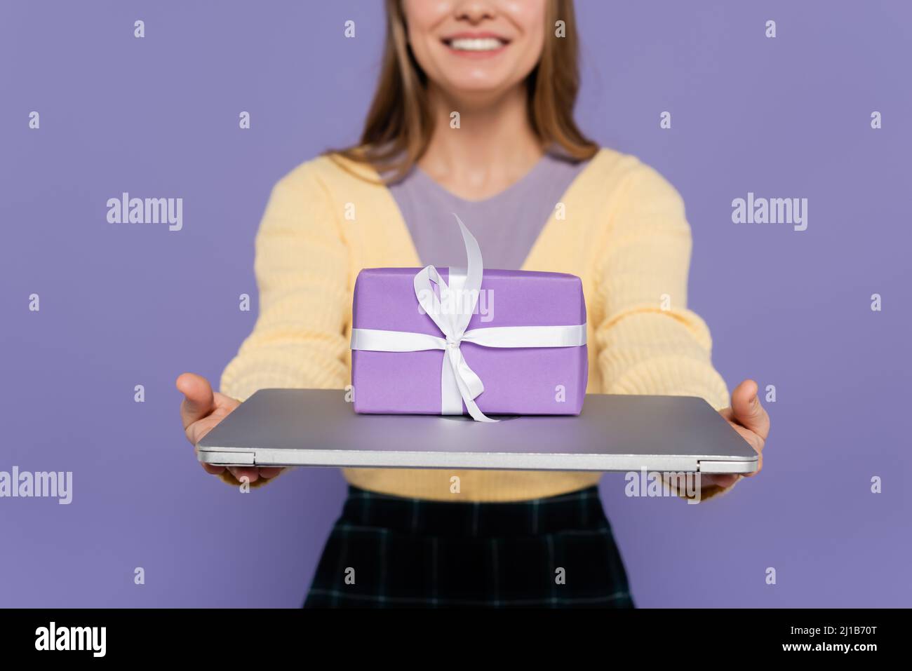 Beschnittene Ansicht der glücklichen jungen Frau mit Laptop mit Geschenkbox isoliert auf lila Stockfoto