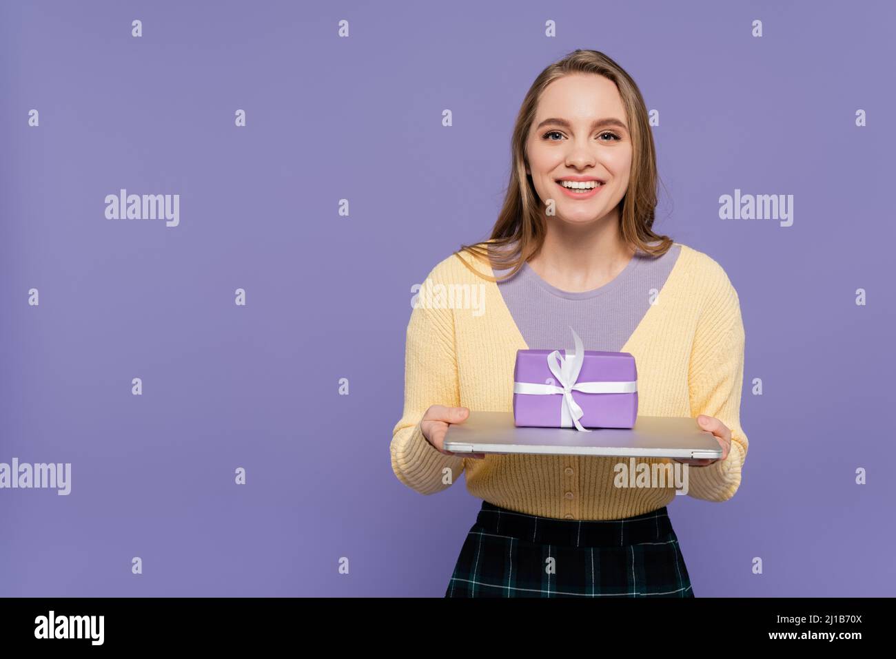 Glückliche junge Frau hält Laptop mit Geschenkbox isoliert auf lila Stockfoto