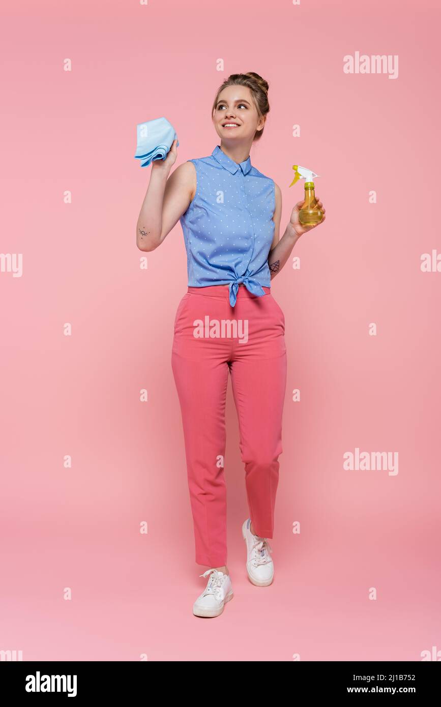 Die ganze Länge der glücklichen und tätowierten Frau hält blauen Lappen und Sprühflasche auf rosa Stockfoto