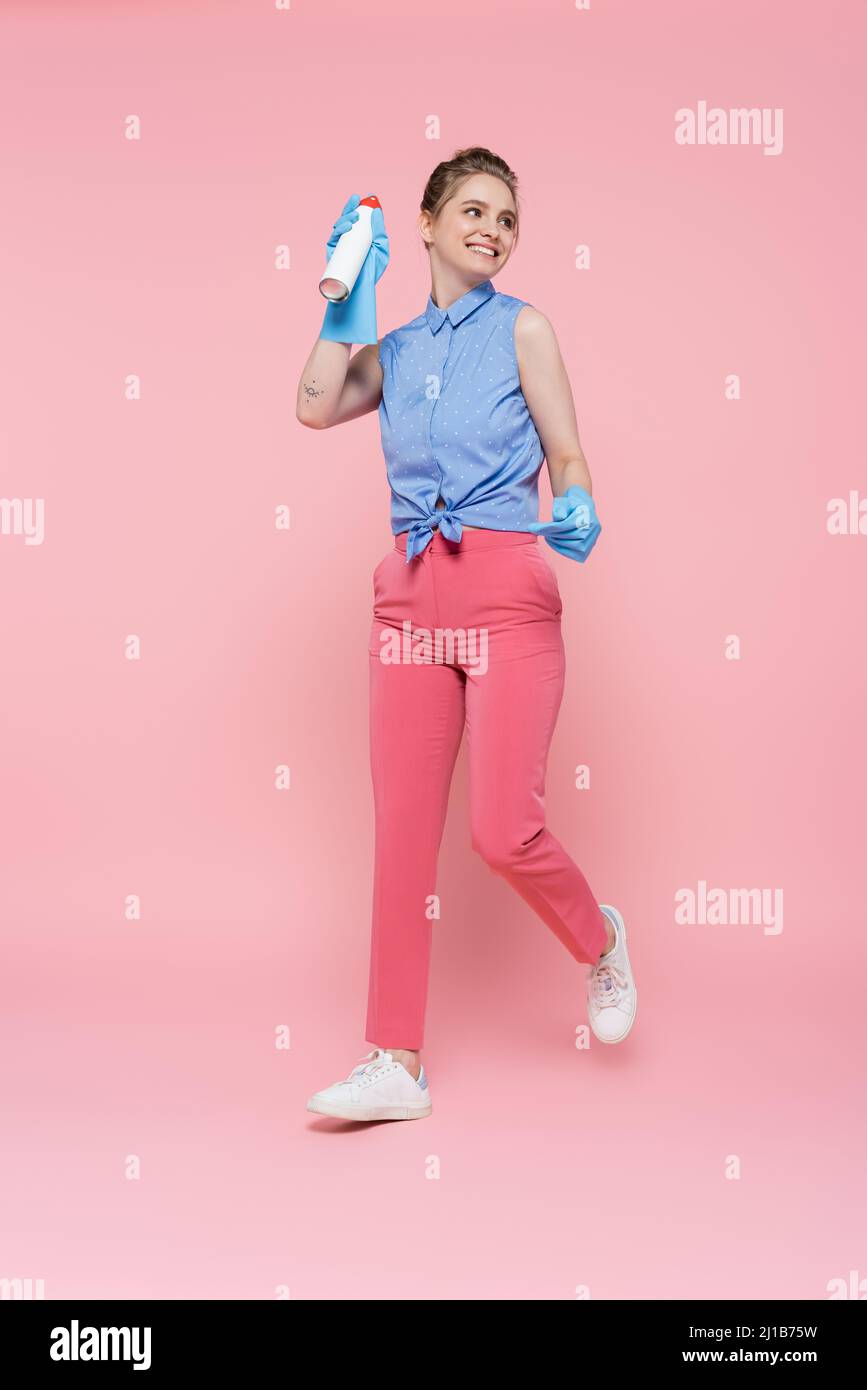 Die ganze Länge der glücklichen jungen Frau in Gummihandschuhen hält Lufterfrischer Flasche auf rosa Stockfoto