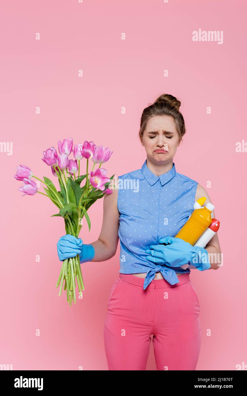 Unzufrieden junge Frau in Gummihandschuhen mit Flaschen mit Waschmittel und Tulpen isoliert auf rosa Stockfoto