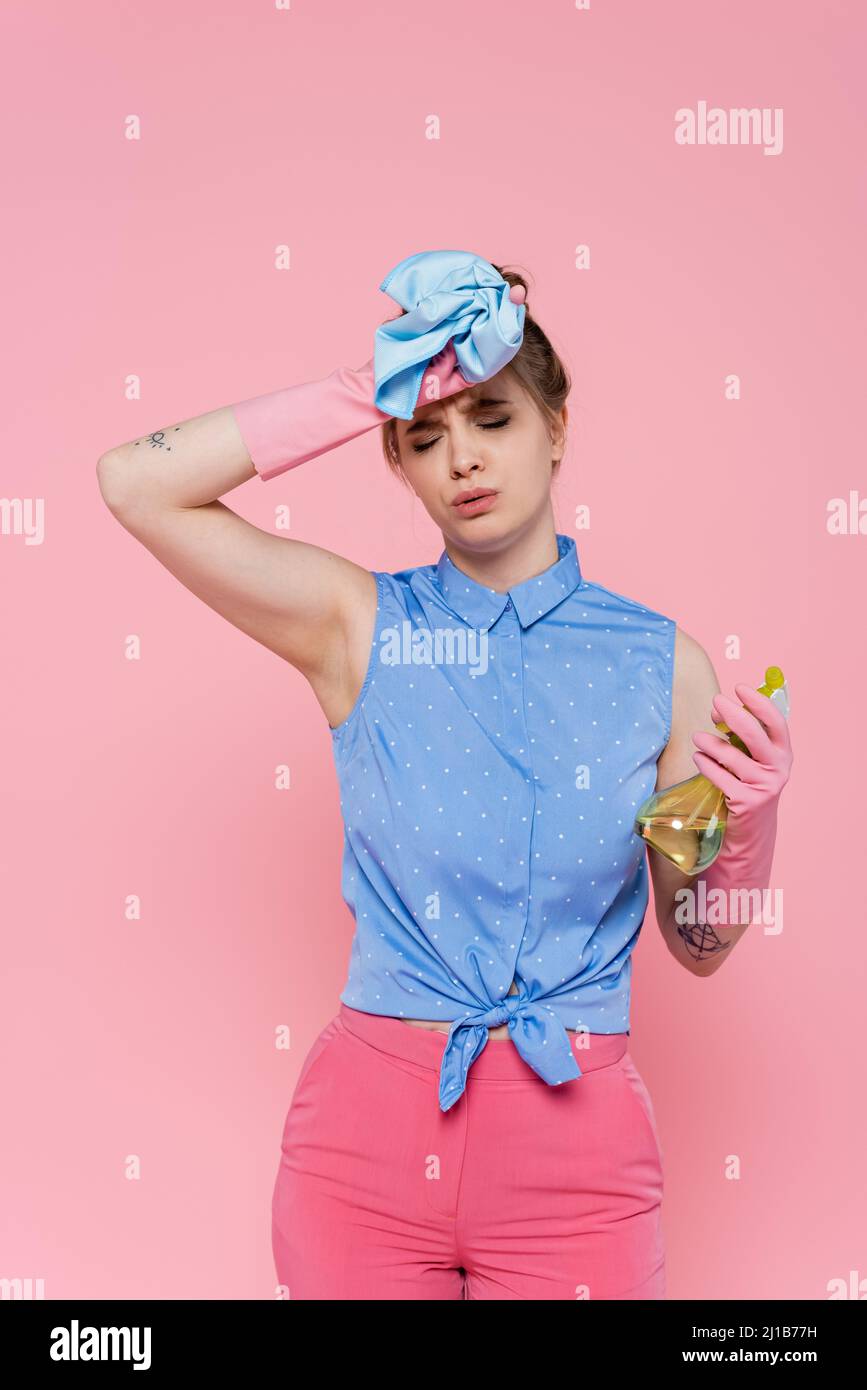 Müde junge Frau in Gummihandschuhen hält blauen Lappen und Sprühflasche isoliert auf rosa Stockfoto