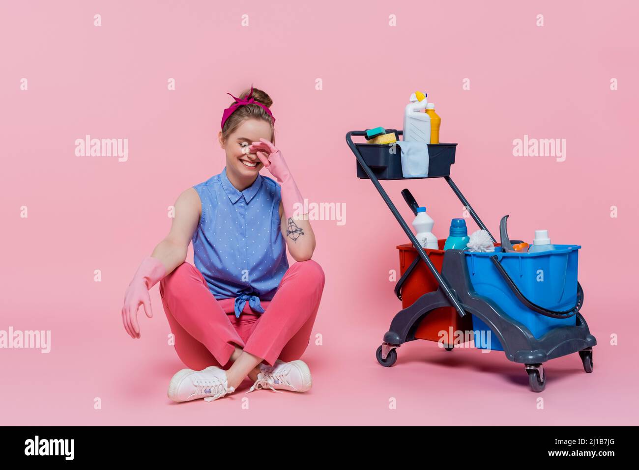 Die ganze Länge der lächelnden jungen Frau in Gummihandschuhen sitzt neben dem Haushaltswagen mit Reinigungsutensilien auf rosa Stockfoto