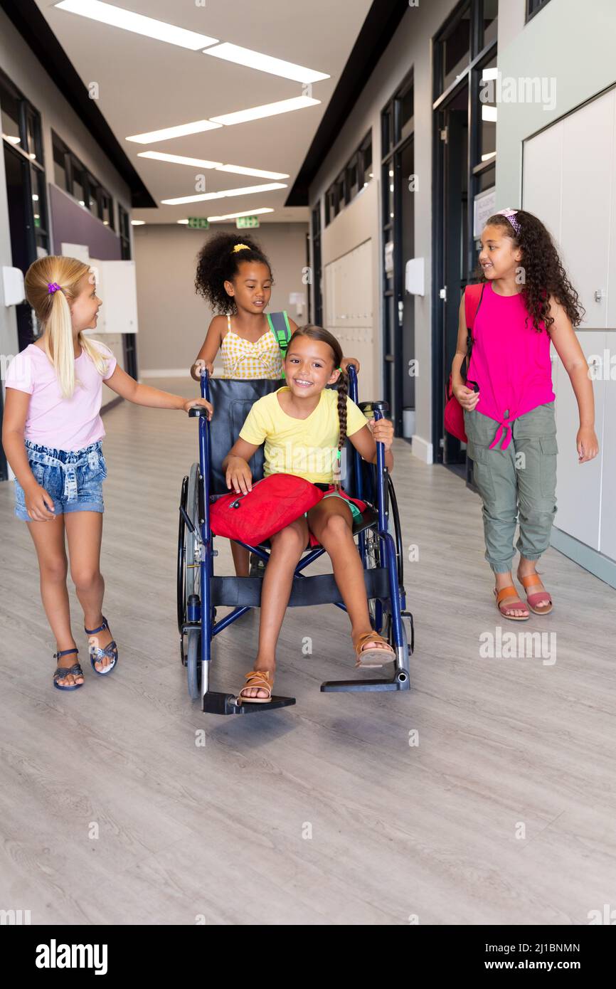 Multirassische Grundschüler, die mit einer Klassenkameradin auf dem Rollstuhl im Korridor laufen Stockfoto