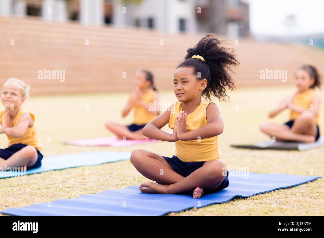 Multirassische Grundschüler mit zusammengeklemmten Händen sitzen auf einer Yogamatte auf dem Schulgelände Stockfoto