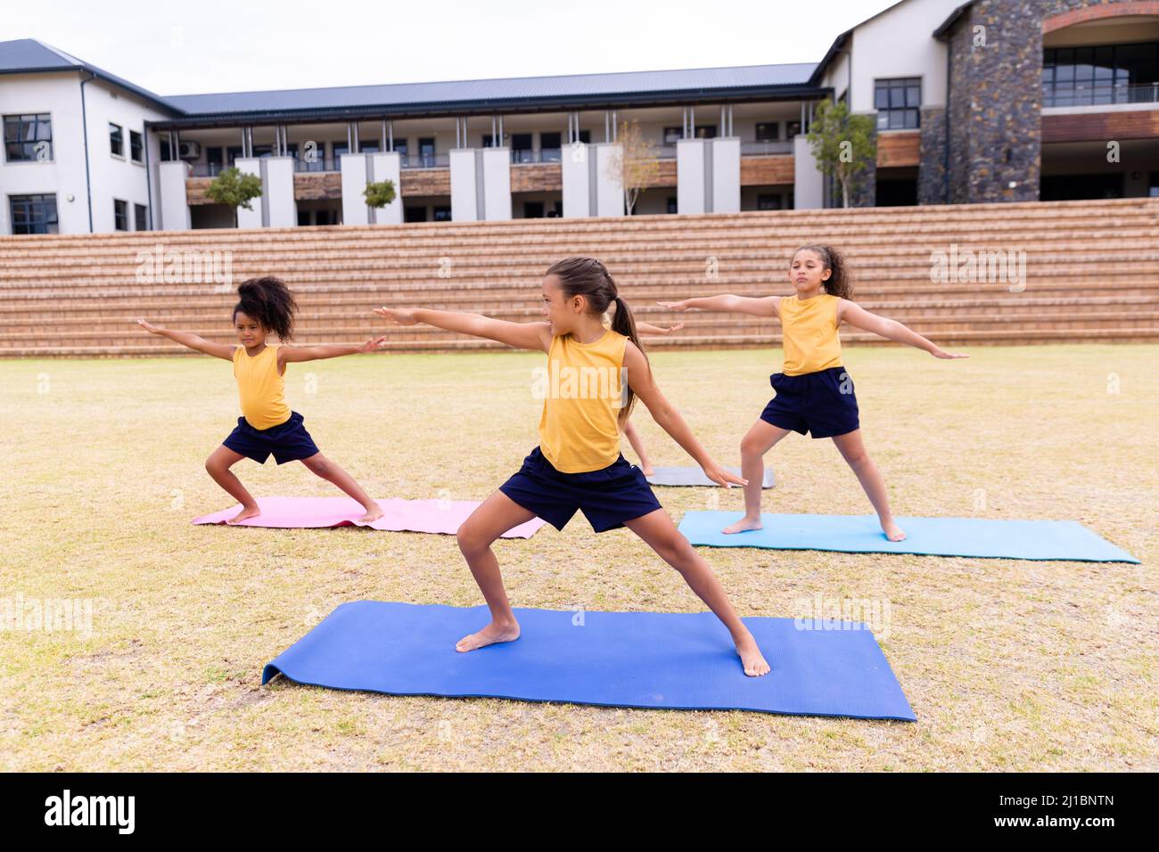 Multirassische Grundschüler tun Krieger 2 Pose beim Training auf Yoga-Matte auf dem Boden Stockfoto