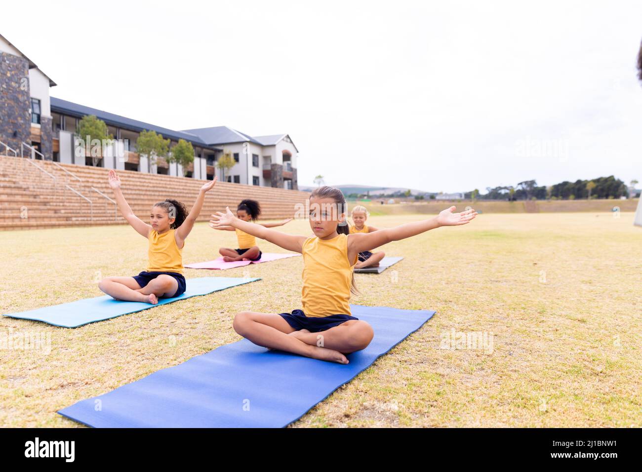 Multirassische Grundschüler mit ausgestreckten Armen trainieren auf einer Yogamatte gegen den Himmel Stockfoto