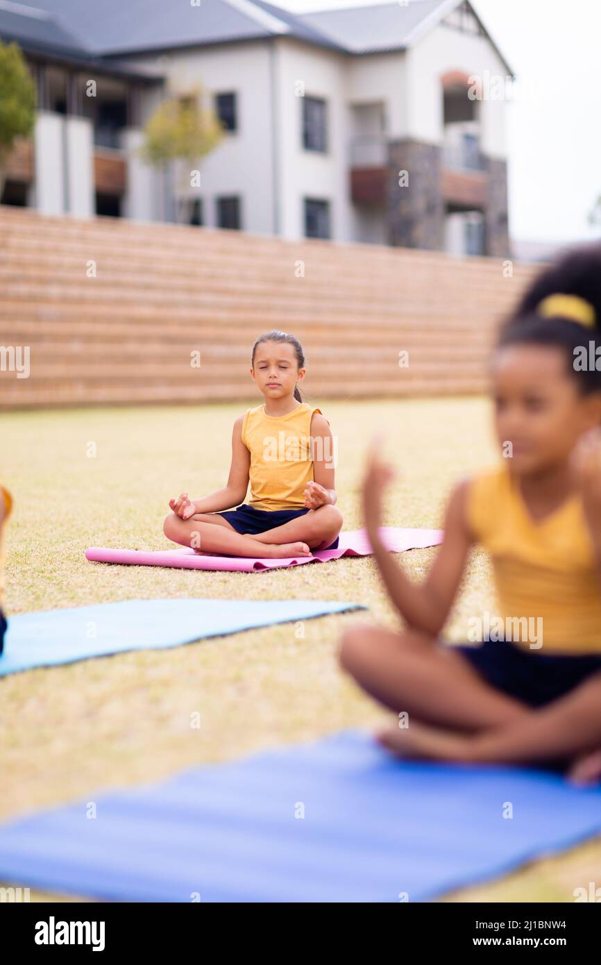 Multirassische Grundschüler meditieren, während sie auf einer Yogamatte gegen die Schule sitzen Stockfoto