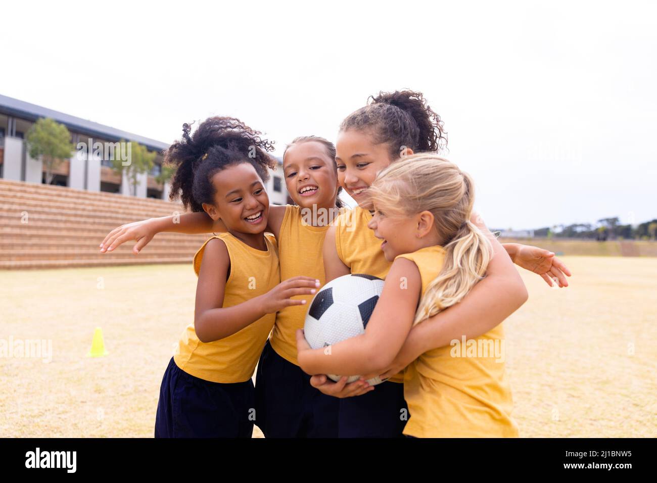 Fröhliche multirassische Grundschüler mit Fußball umarmen, während sie auf dem Boden stehen Stockfoto