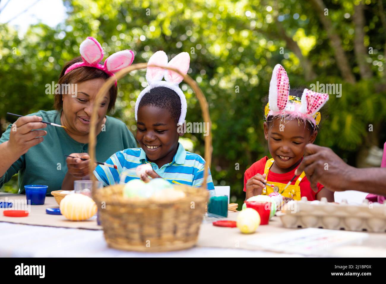 Lächelnde afroamerikanische Geschwister und Großmutter tragen Hasenohren, während sie an ostern Eier malen Stockfoto
