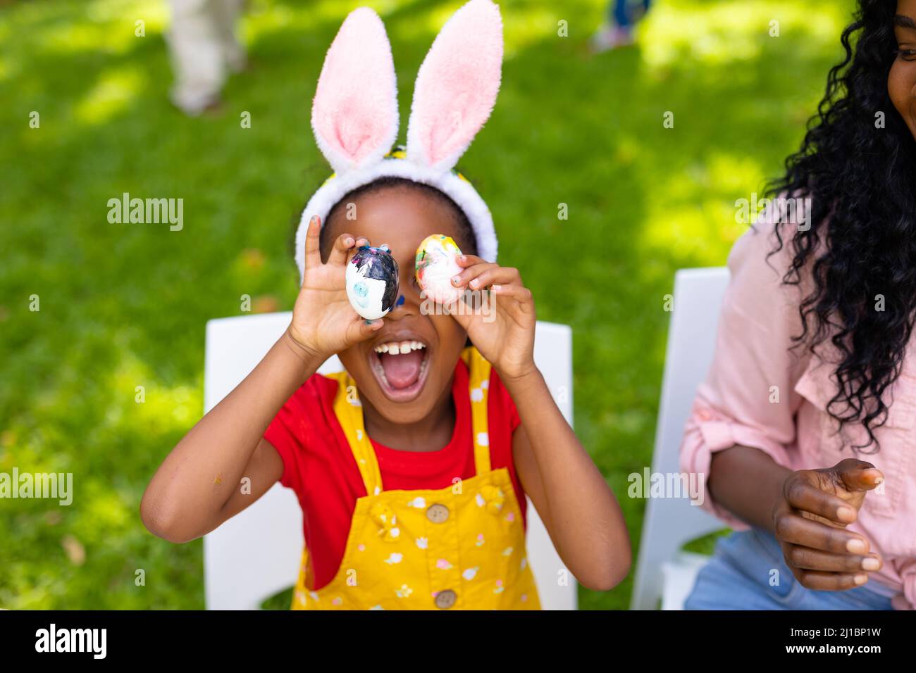 Fröhliche afroamerikanische Mädchen und Mutter spielen mit ostereiern, während die Familie im Hintergrund Stockfoto