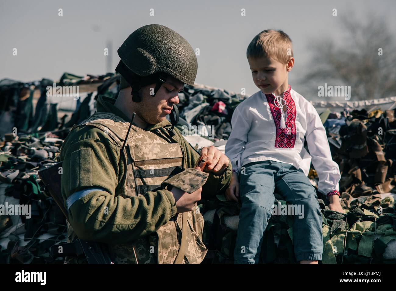 Der Junge im bestickten Hemd sitzt auf Sandsäcken am Checkpoint in der Nähe des ukrainischen Territorialverteidigungskriegers. Konzept der russischen Militärinvasion in Ukra Stockfoto