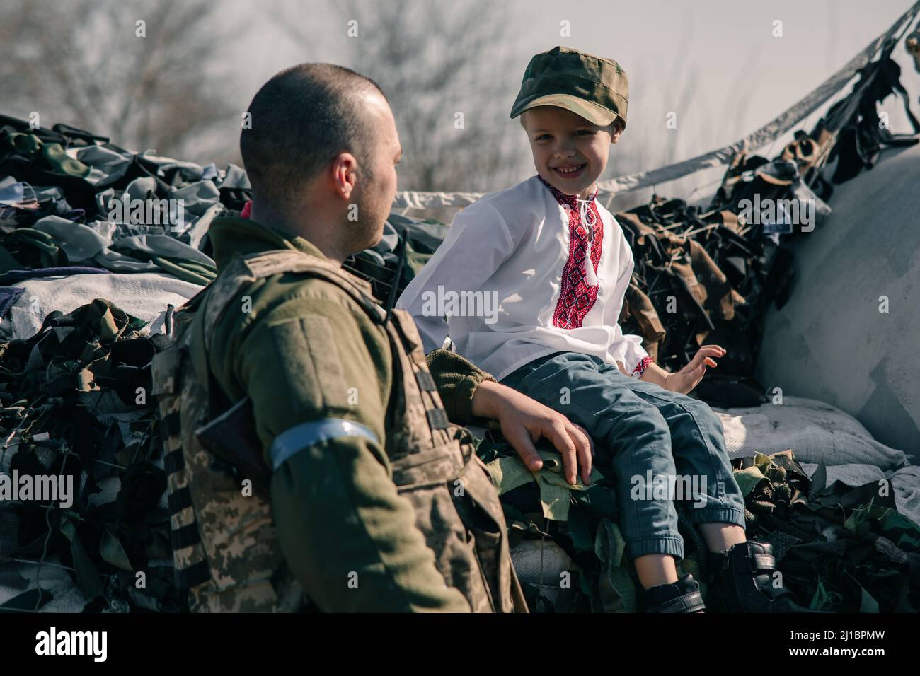 Der Junge im bestickten Hemd sitzt auf Sandsäcken am Checkpoint in der Nähe des ukrainischen Territorialverteidigungskriegers. Konzept der russischen Militärinvasion in Ukra Stockfoto