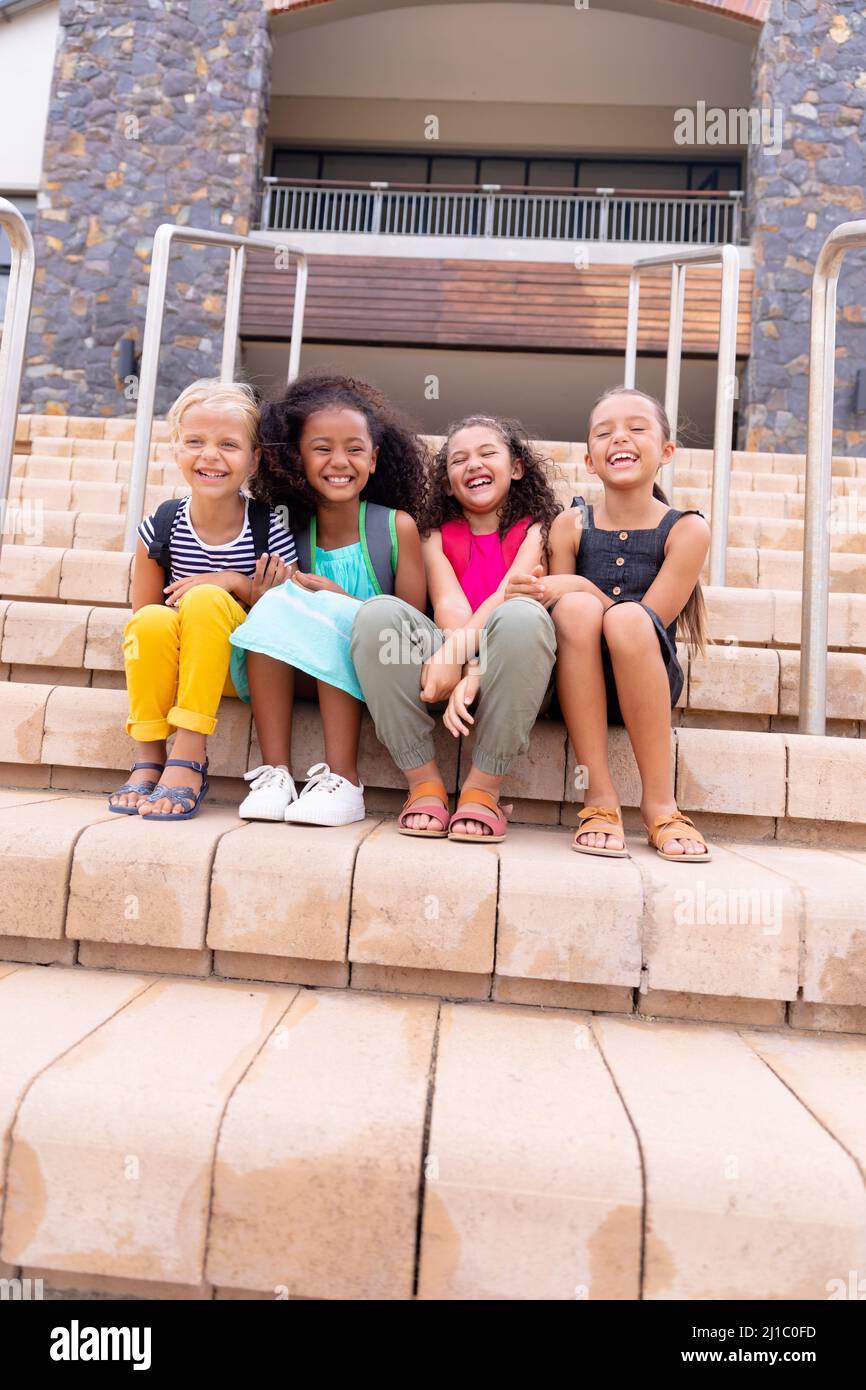 Die ganze Länge der multirassischen fröhlichen Grundschüler sitzen auf Stufen gegen das Schulgebäude Stockfoto