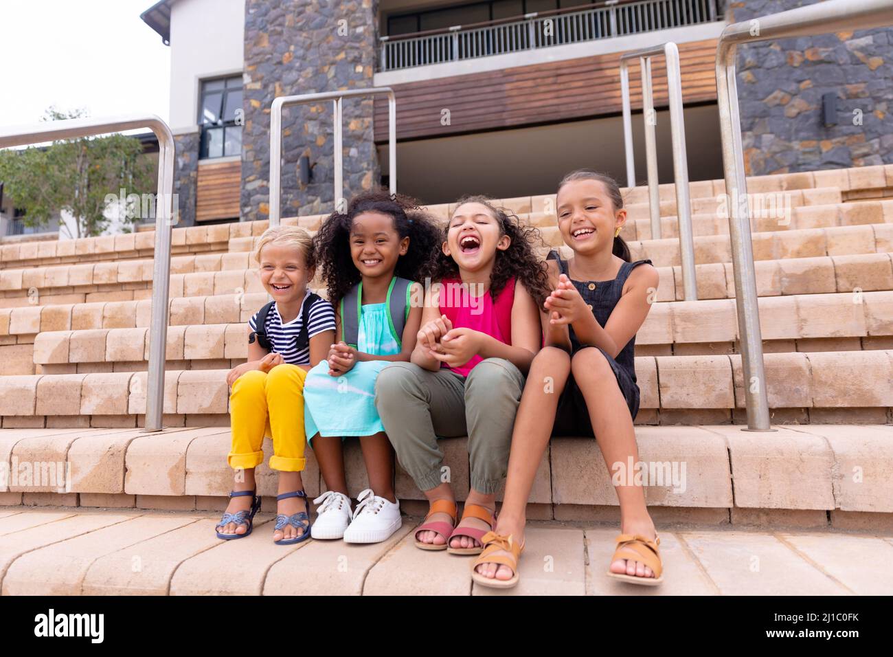 Multirassische fröhliche Grundschüler, die auf Stufen gegen das Schulgebäude sitzen Stockfoto